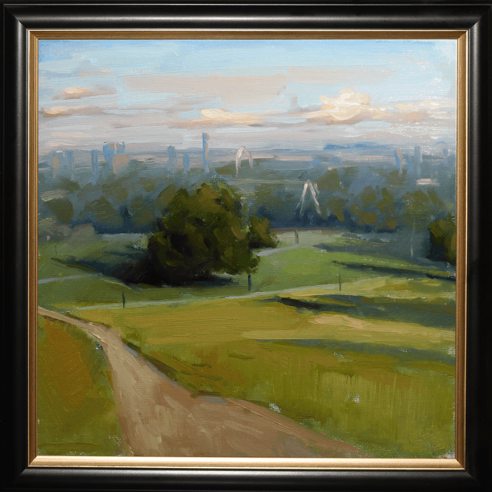 A framed landscape oil painting featuring a green field with a dirt path, trees, and a distant city skyline under a cloudy sky, enclosed in a black frame with a gold inner border.