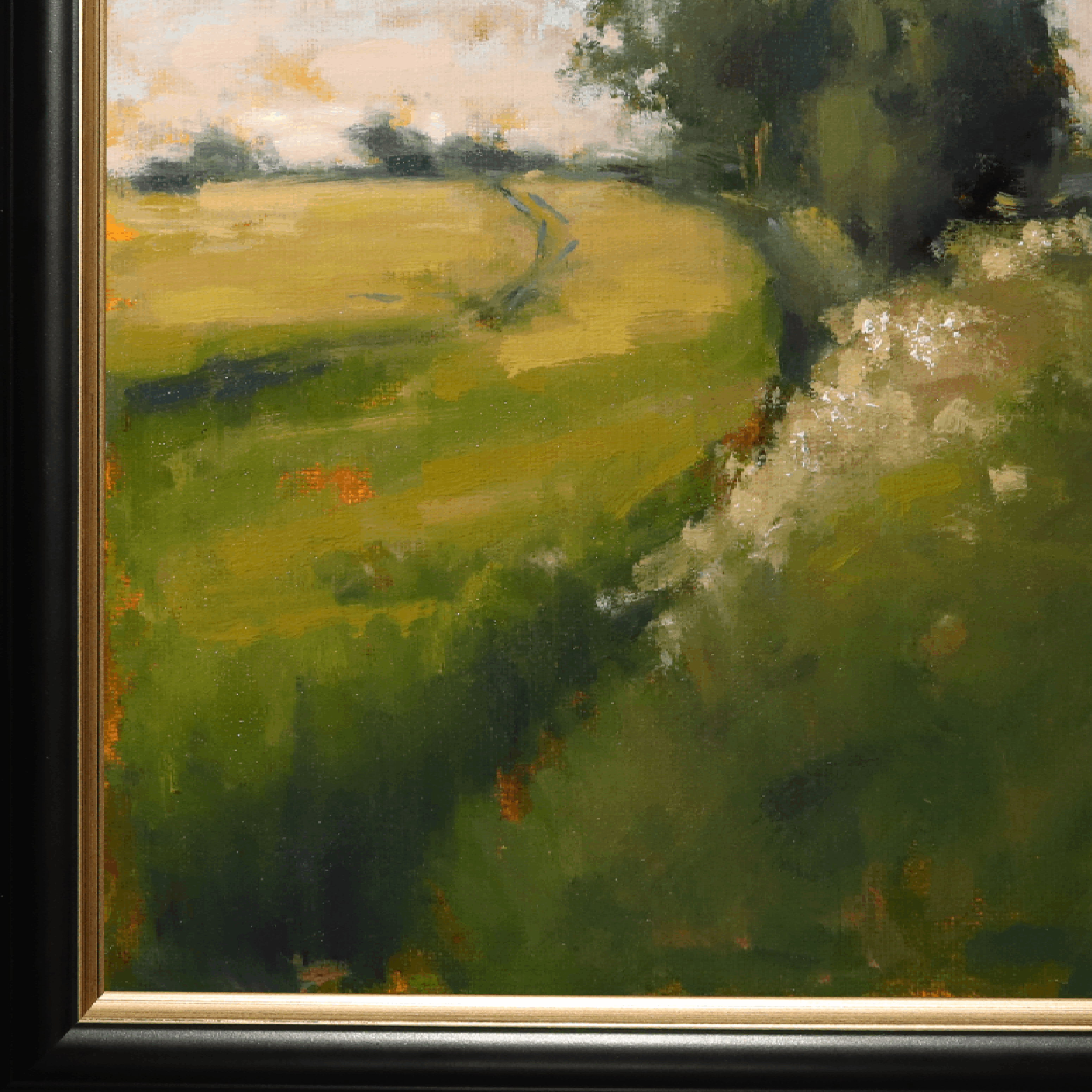 A close-up of a framed landscape painting featuring a green field with a winding path, dark trees on the right, and a light sky. The painting is bordered by a black frame with a gold inner edge.