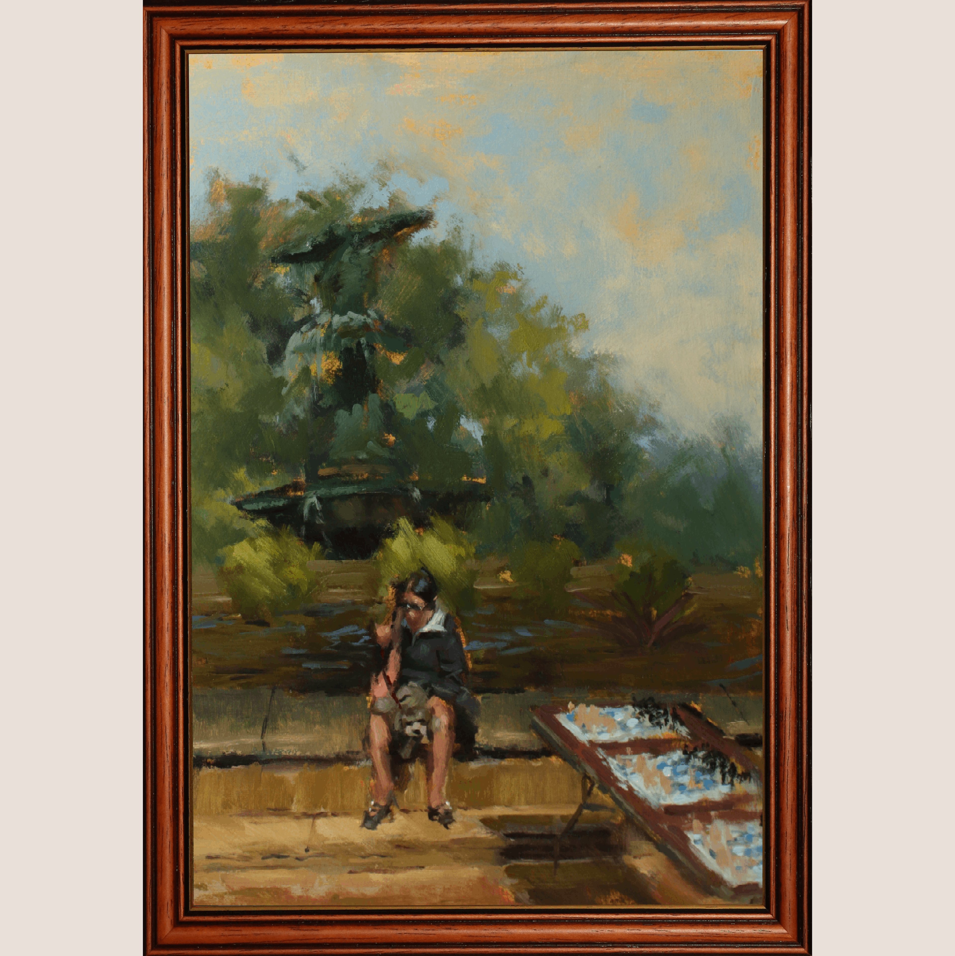 A framed oil painting depicting a seated figure near a table in an outdoor garden setting, enclosed in a warm brown wooden frame against a light beige background.