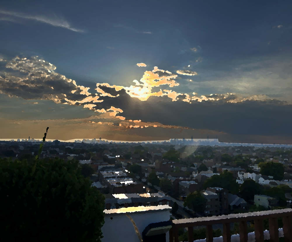 A scenic sunset over a suburban city skyline, with dramatic clouds illuminated by the setting sun and sunbeams breaking through. The foreground features dark green trees and a wooden railing, looking out over a dense residential neighborhood.