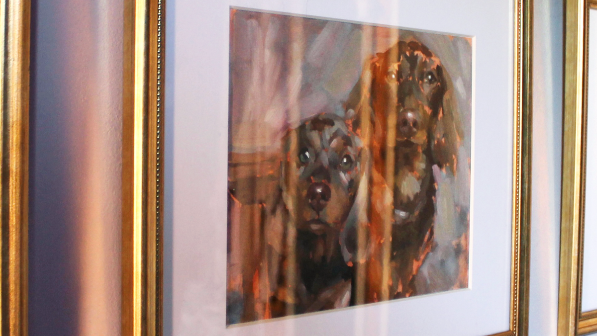 A framed painting of two dogs, likely a Cocker Spaniel, rendered in warm brown, orange, and black tones with a white mat and ornate gold frame.