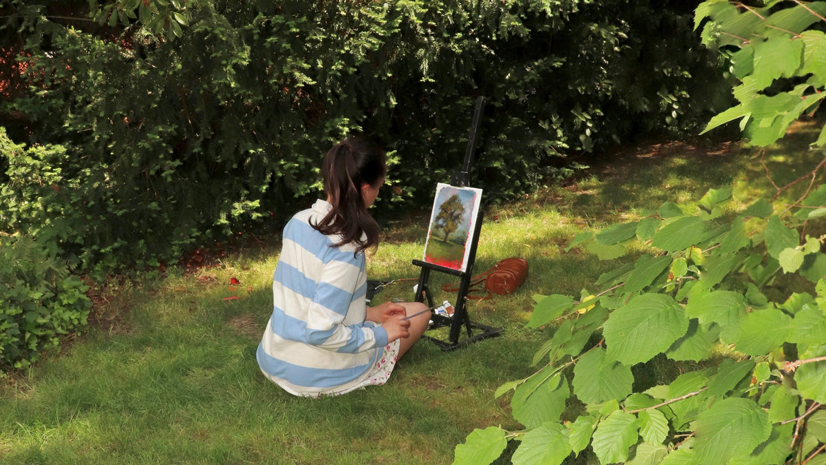 A woman sits on the grass in a garden, painting on an easel. She is wearing a light blue and white striped shirt.