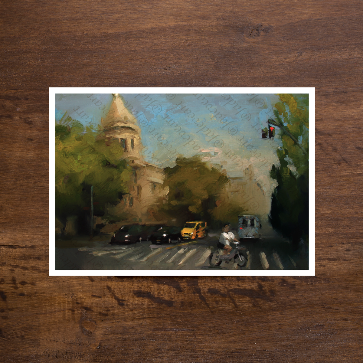 A square art print featuring an impressionistic painting of a city street scene with trees, a domed building, cars, and a cyclist, displayed on a dark brown wooden surface.