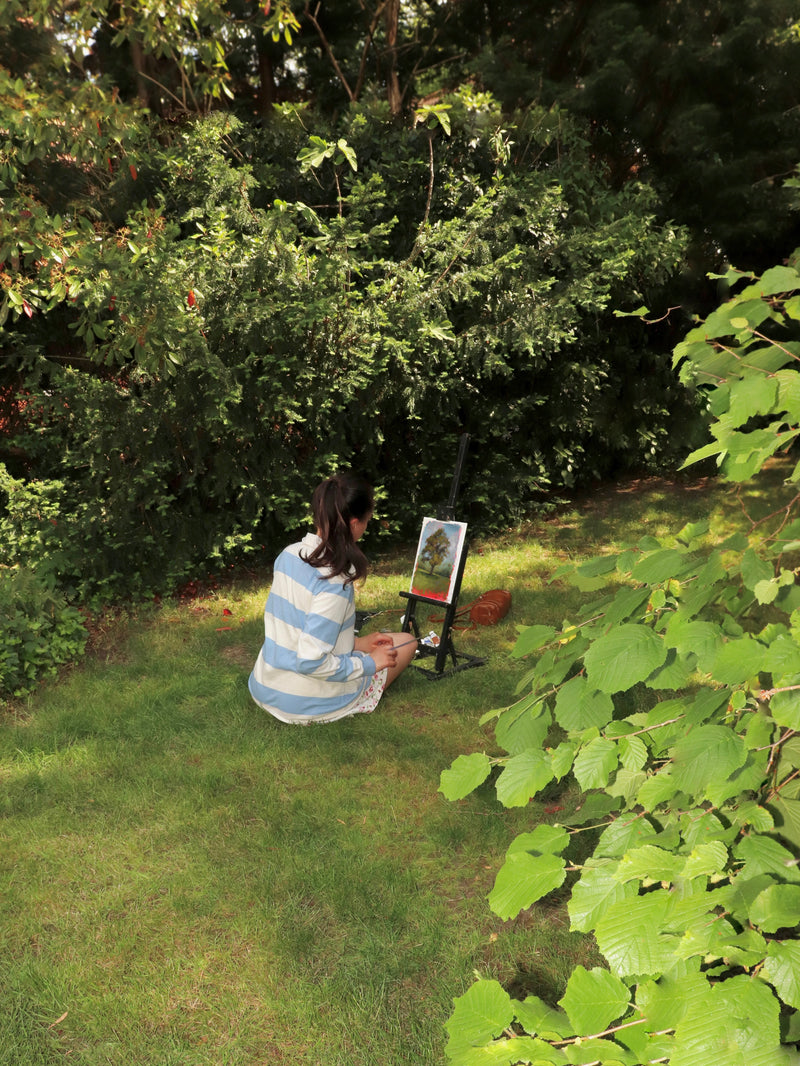 A person sits on grass in a wooded outdoor setting, facing an easel with a painting in progress. The person is wearing a blue and white striped long-sleeve top.