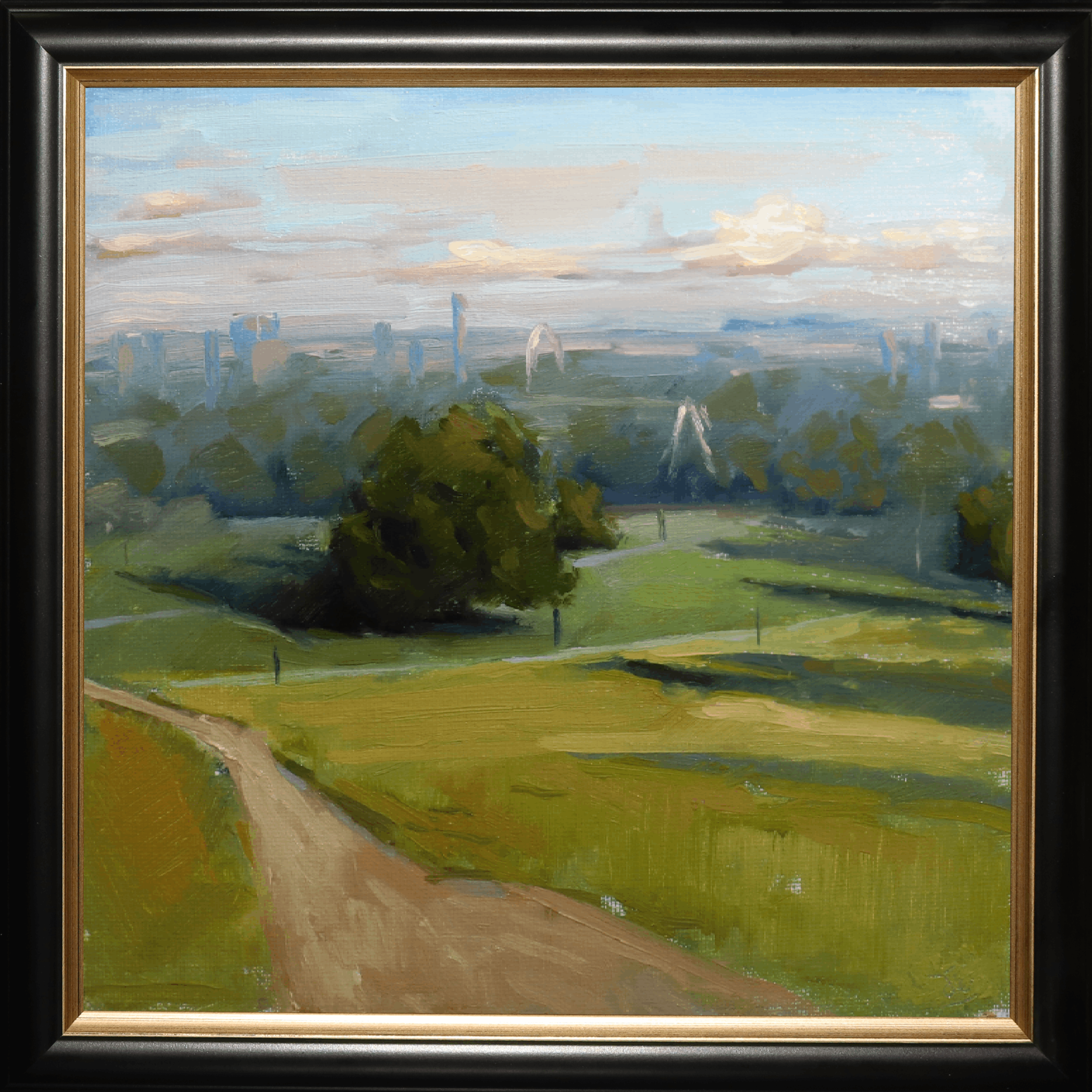A framed landscape oil painting featuring a green field with a dirt path leading to a cluster of trees, with a distant city skyline under a blue and white sky. The painting is housed in a black frame with a gold inner border.