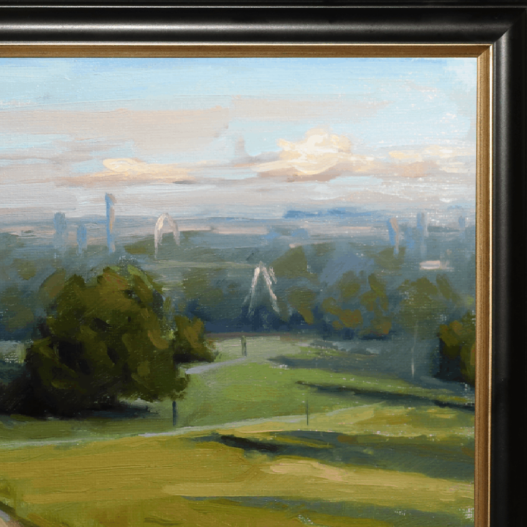 A close-up of a framed landscape painting showing green fields, trees, distant buildings, and a pale blue sky with soft clouds. The painting is bordered by a dark frame with a gold inner edge.