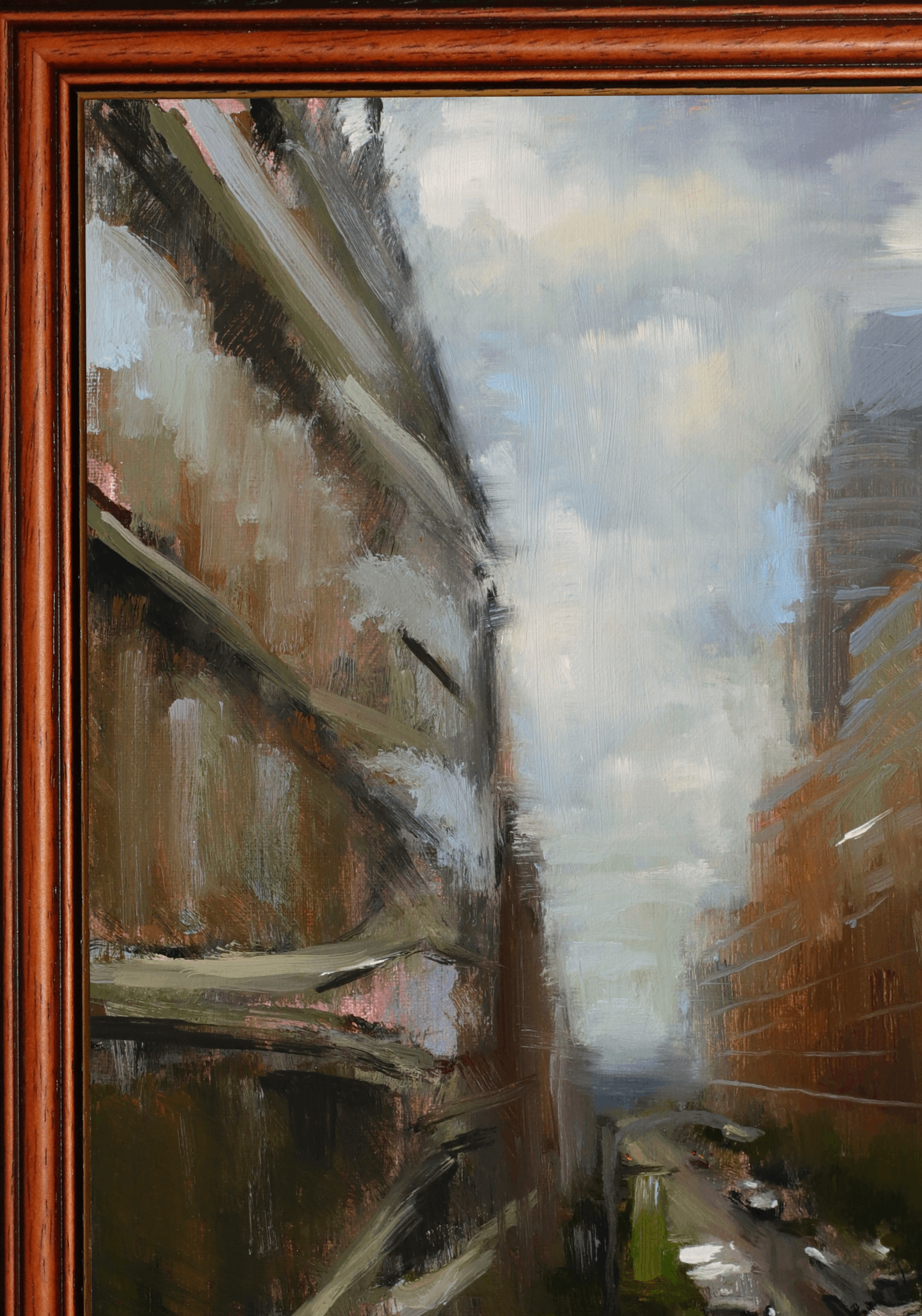 An impressionistic oil painting of a narrow city street flanked by tall buildings under a cloudy sky, presented in a warm brown wooden frame.